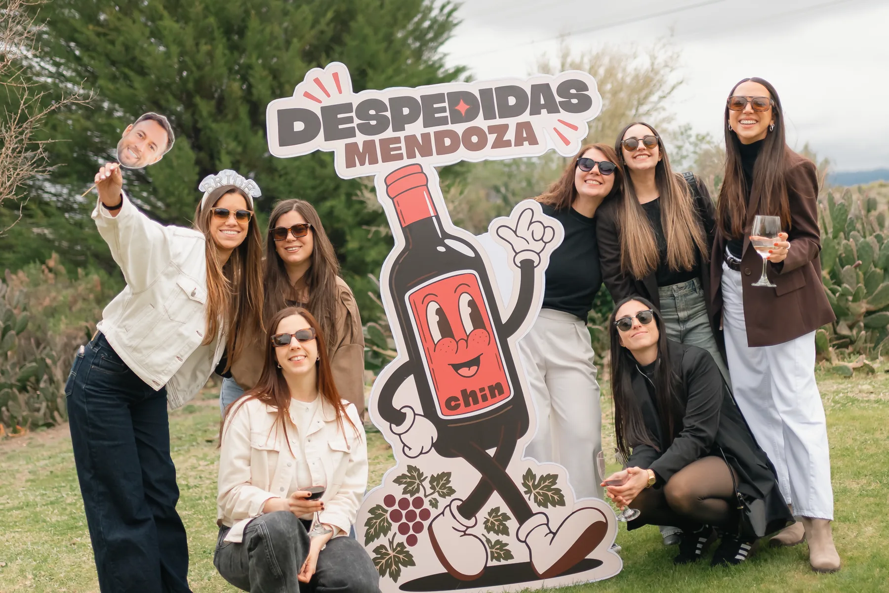 Grupo de amigas con copas junto a la gigantografía de Chin y el cartel de Despedidas Mendoza, al aire libre.